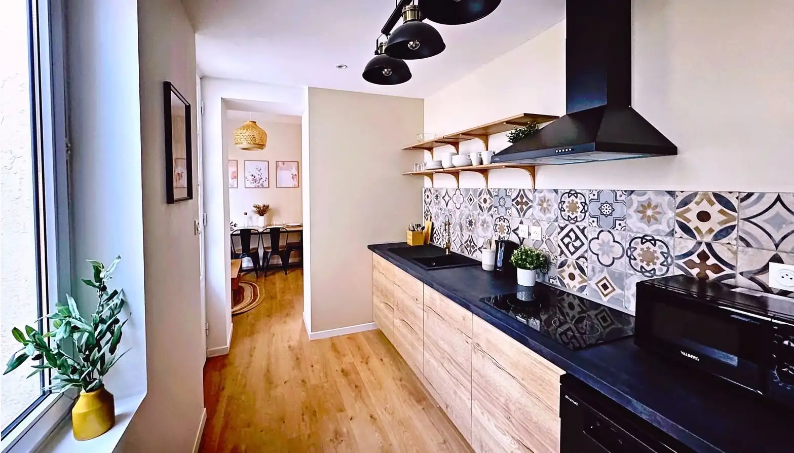 Cuisine fonctionnelle et lumineuse avec murs couleur lin, crédence en carreaux de ciment, plan de travail en granit noir, frigo pour 4 personnes, et suspension luminaire atelier.