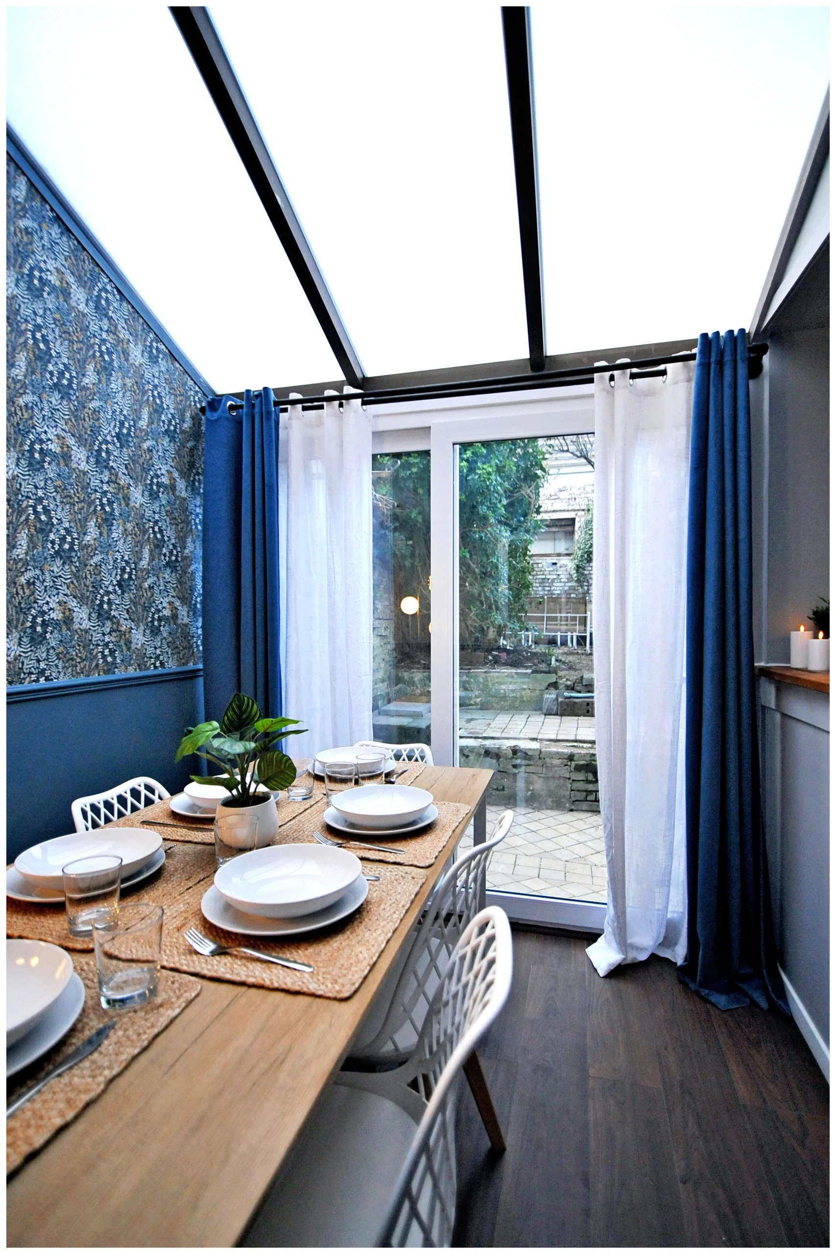 Salle à manger rénovée en colocation, sous verrière, avec une grande table pour six, murs bleu moderne, tapisserie maison bourgeoise et luminaire doré tendance 2025.