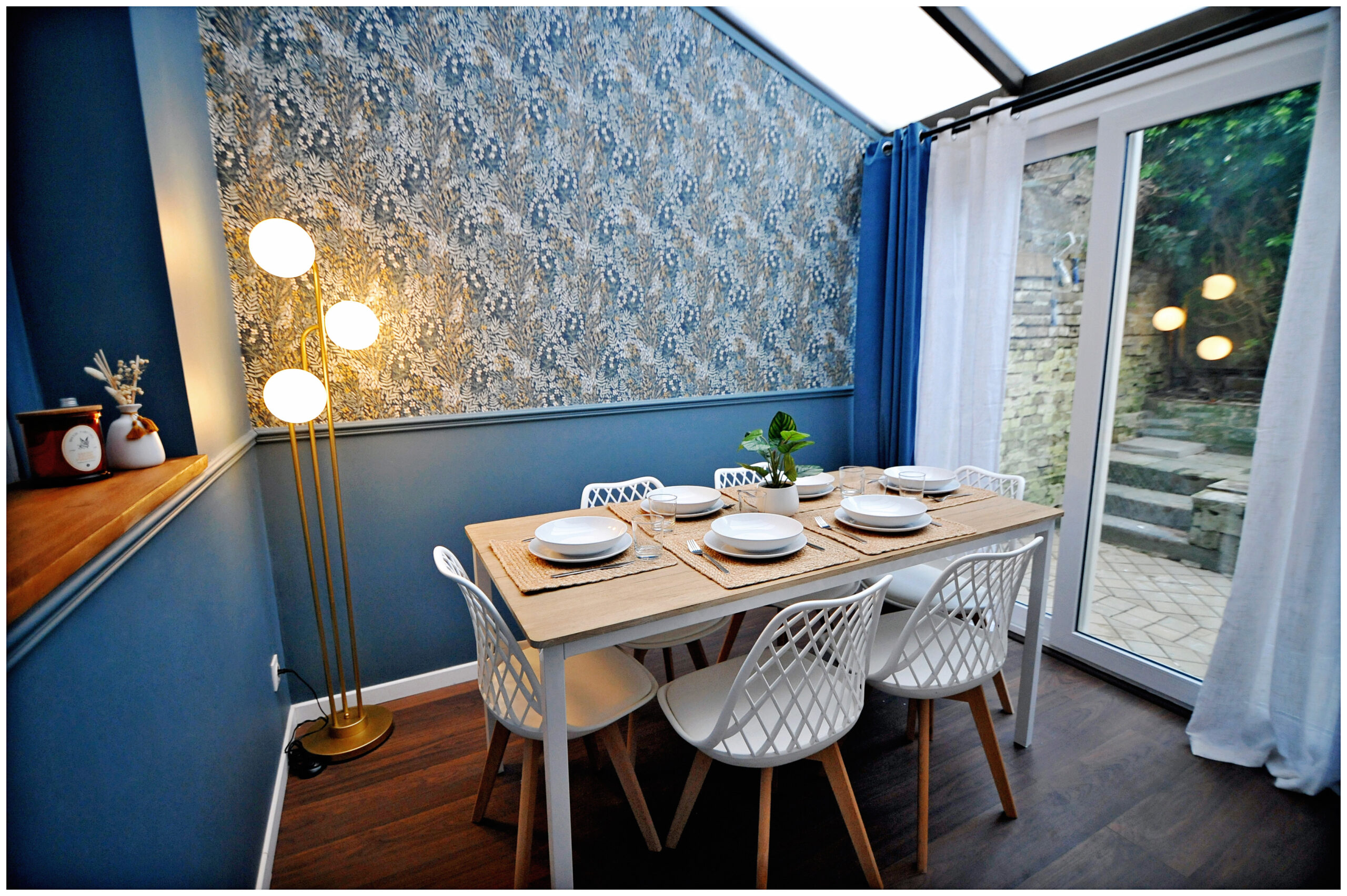 Salle à manger rénovée en colocation, sous verrière, avec une grande table pour six, murs bleu moderne, tapisserie maison bourgeoise et luminaire doré tendance 2025.